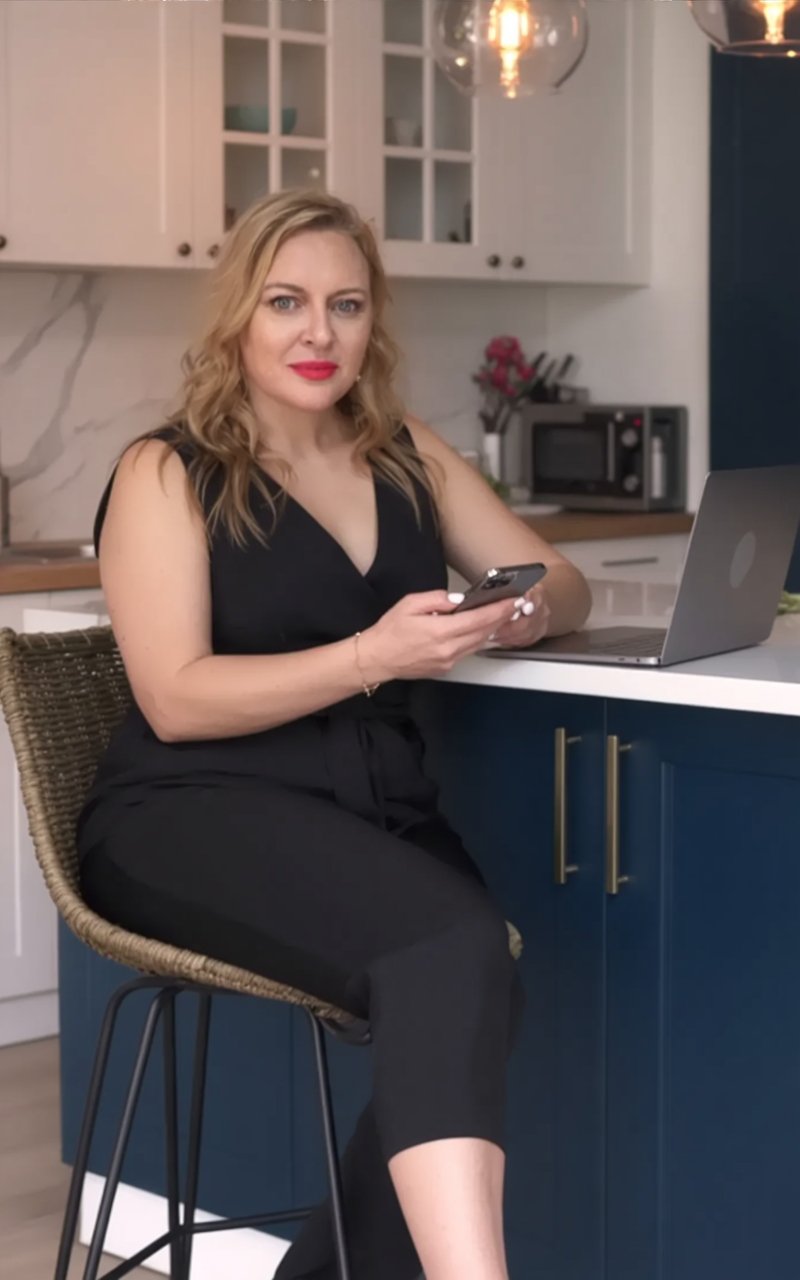 Andrea Florescu working on a laptop at a kitchen counter with her phone in hand, illustrating a flexible, modern approach to running a consulting business