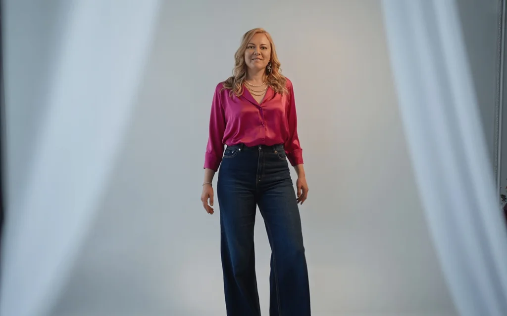 Founder in a fuchsia blouse and wide-leg jeans stepping forward between drawn curtains — an invitation to step into alignment