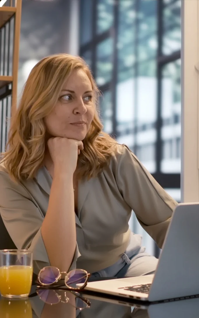 Strategist seated at a laptop with hand on chin, gazing thoughtfully out a window — grounded clarity in the middle of complex work