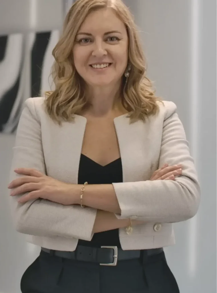 Andrea Florescu standing confidently with arms crossed, wearing a light blazer and smiling in a modern professional office setting.